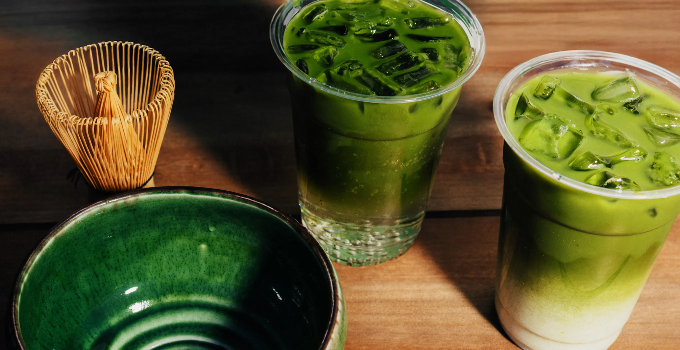 Matcha drinks and tea utensils lined up on a wooden table. On the left is a traditional bamboo chasen (tea whisk) and a dark green matcha bowl, and on the right are two matcha iced drinks with ice in transparent cups.